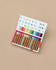 An open box of Kitpas Crayons - 12 Colors rests on a light brown surface, displaying the non-toxic crayons neatly arranged with a color guide chart printed inside the lid.