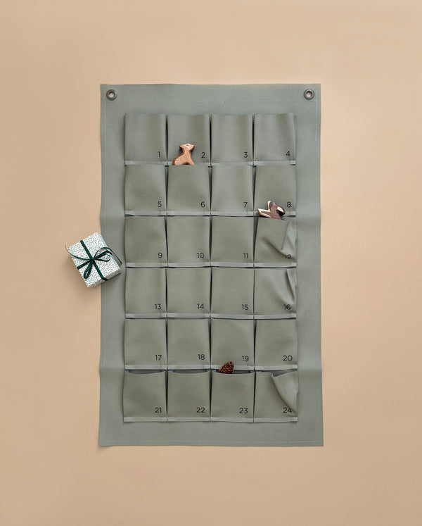 The Gathre Small Advent Calendar - Grove, featuring 24 numbered gift pockets in a minimalist fabric design, is displayed on a beige wall for an ideal Christmas countdown. Three of its pockets contain charming small toys: a cloth bunny, a peg doll, and a fabric pinecone. Next to it lies a petite silver gift box wrapped with a black ribbon.