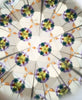 The Konges Sloejd Kaleidoscope - Lucia features symmetrical green, yellow, purple, and orange shapes with geometric lines creating an intricate, repeating design on a light background embellished with colorful pearls.