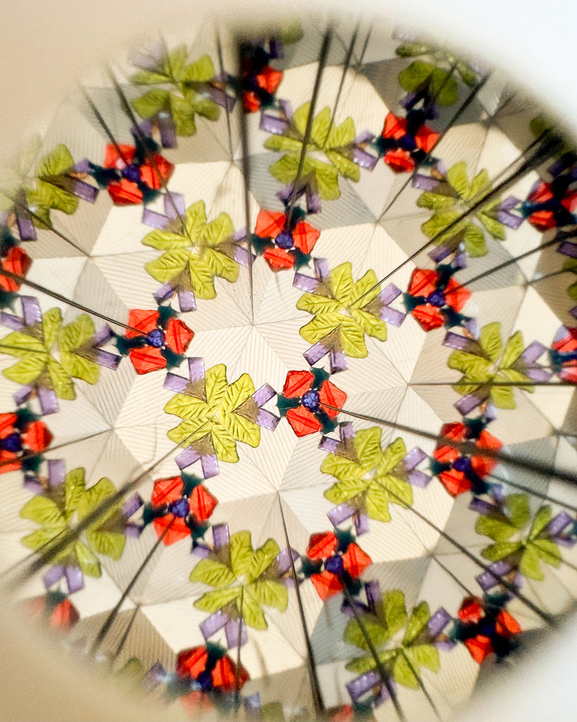 Peer through the Konges Sloejd Kaleidoscope - Like Cats and Dogs to discover a mesmerizing, symmetrical pattern of green, red, and purple shapes—reminiscent of leaves and jewels—arranged in a repeating geometric design, highlighted with vibrant pearl-like accents.