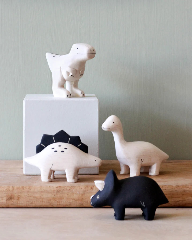 Four stylized dinosaur figurines handcrafted from Albizia wood displayed on a wooden surface against a green wall. The set includes a white T-rex on a white block, a Handmade Tiny Wooden Dinosaurs - Brachiosaurus.