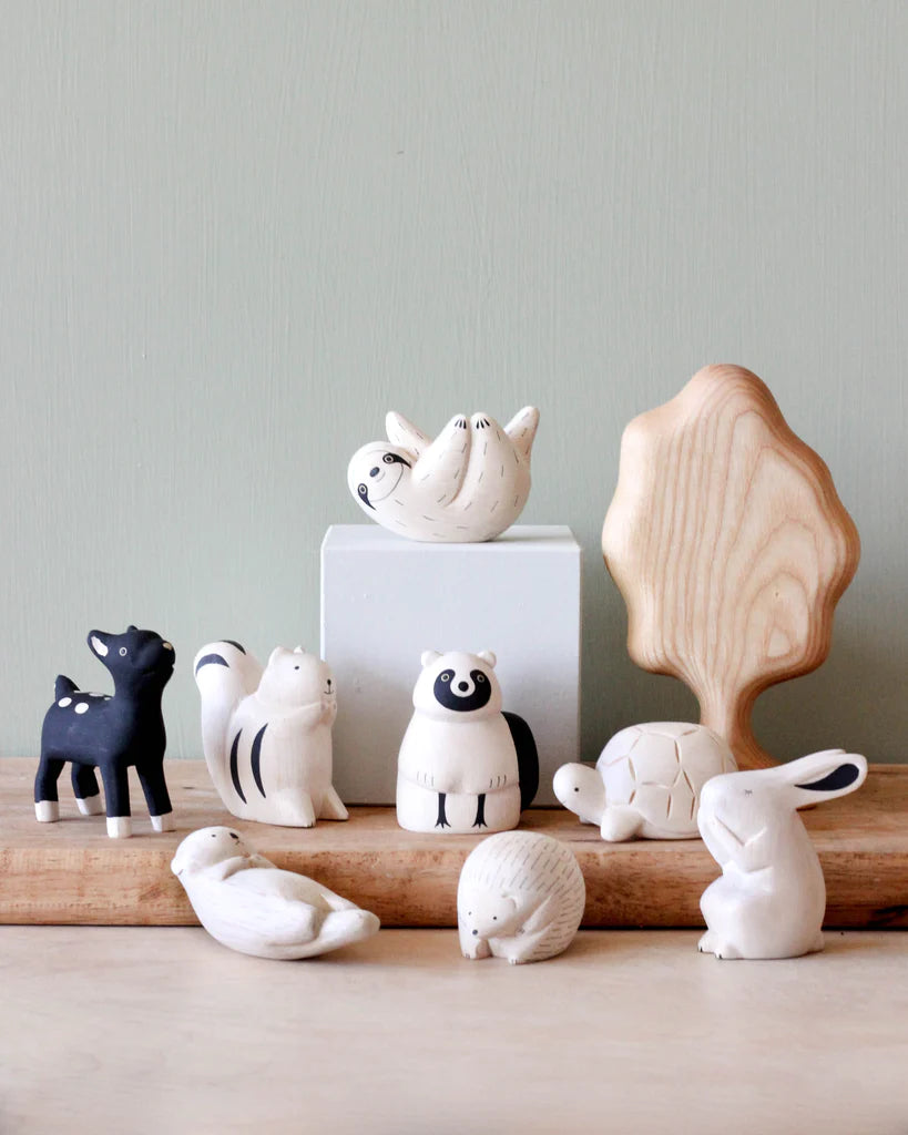 A collection of small, minimalist animal figurines, including a black deer, a white squirrel, a raccoon (Handmade Tiny Wooden Forest Animals - Raccoon), a turtle, a rabbit, and a bird. These handcrafted wooden animals are displayed on wooden surfaces with a light green background and an Albizia wood tree prop.