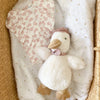 Mon Ami - Colette the Duck Plush Toy, a soft white duckling with a pink floral collar, rests beside a pink leafy-patterned baby outfit on a gold star blanket inside a woven basket.