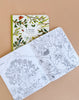 The Moulin Roty Botanist Coloring Book & Stickers, open to detailed flora and fauna pages, reveals trees and animals. Below it rests a closed copy with a leafy bird-illustrated cover on a beige surface.