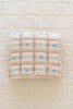 The Organic Cotton Baby Quilt - Meadow, Petal, made in Portugal, features a pink and white plaid with small blue flowers and lace trim. It measures 30x40 inches and rests on a quilted white surface.