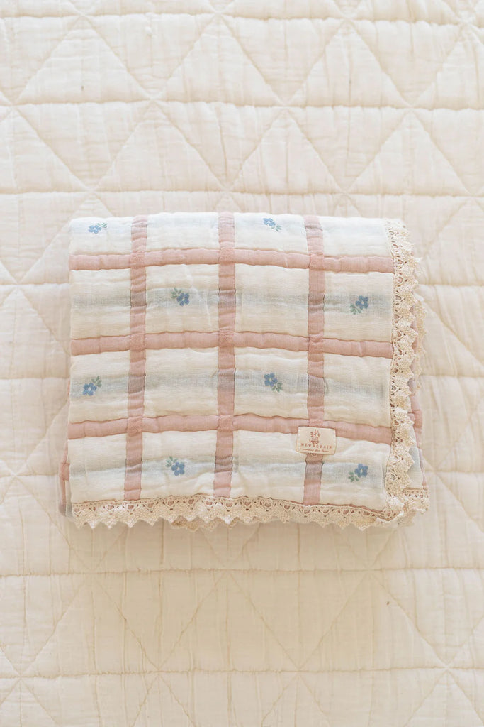 The Organic Cotton Baby Quilt - Meadow, Petal, made in Portugal, features a pink and white plaid with small blue flowers and lace trim. It measures 30x40 inches and rests on a quilted white surface.