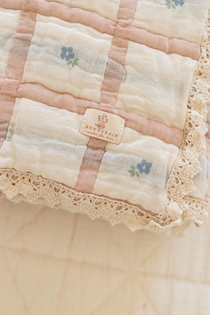 A close-up of the Organic Cotton Baby Quilt - Meadow, Petal: a soft, pastel quilt with delicate lace edges, blue floral patterns, and a "NEW BRAIN" tag. This cozy 30x40 inch vintage-style quilt is made in Portugal from organic cotton.