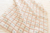 The Organic Cotton Baby Quilt - Meadow, Petal, made in Portugal with pastel pink and beige checks and lace edging, is spread out atop a matching patterned bedspread.