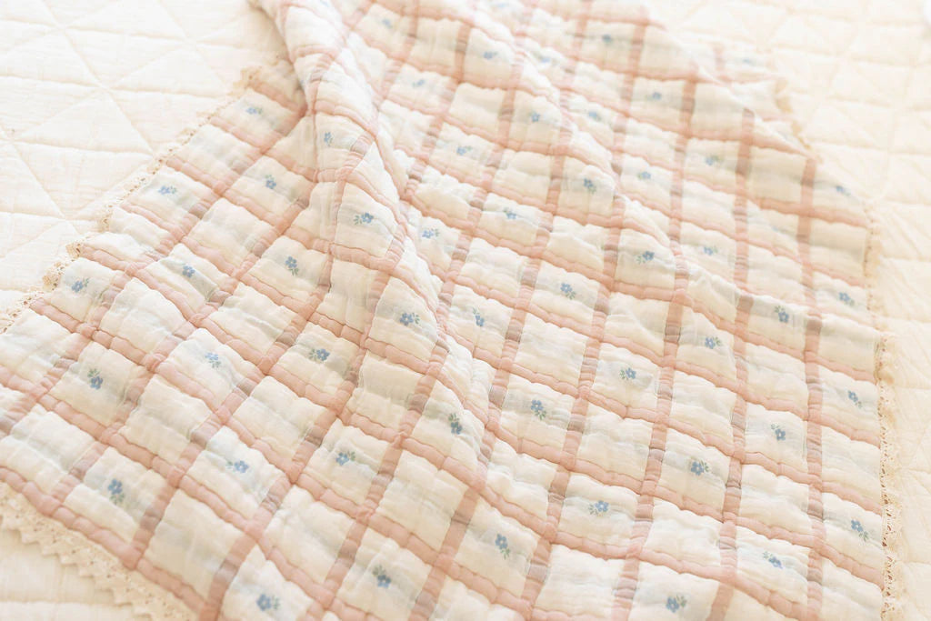 The Organic Cotton Baby Quilt - Meadow, Petal, made in Portugal with pastel pink and beige checks and lace edging, is spread out atop a matching patterned bedspread.