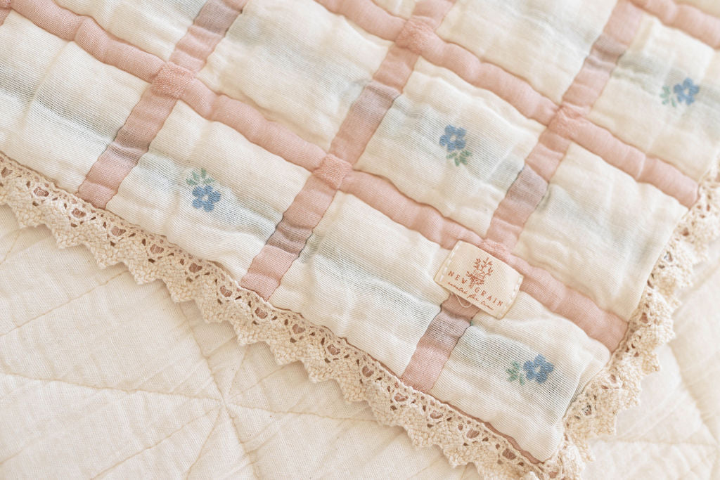 A close-up of the Organic Cotton Baby Quilt - Meadow, Petal shows its soft, quilted organic cotton with a pastel checkered pattern, blue floral accents, beige lace trim, and a 30x40 inch size with a fabric label sewn in one corner.