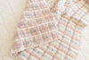 The Organic Cotton Baby Quilt - Meadow, Petal features a soft pink and white plaid pattern with blue flower accents and delicate lace trim, adding a gentle touch to a light-colored bed.