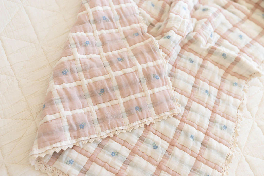 The Organic Cotton Baby Quilt - Meadow, Petal features a soft pink and white plaid pattern with blue flower accents and delicate lace trim, adding a gentle touch to a light-colored bed.