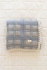 The Organic Cotton Baby Quilt - French Plaid, Bluebelle is a 30x40-inch folded blue and gray plaid quilt with lace trim, made in Portugal, featuring a small fabric label on the front and resting on a white quilted surface.