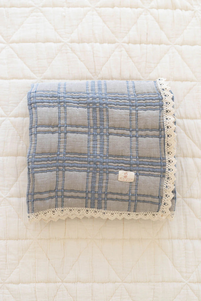 The Organic Cotton Baby Quilt - French Plaid, Bluebelle is a 30x40-inch folded blue and gray plaid quilt with lace trim, made in Portugal, featuring a small fabric label on the front and resting on a white quilted surface.