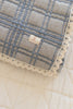 The Organic Cotton Baby Quilt - French Plaid, Bluebelle features a folded light blue and gray checkered pattern with cream lace trim and "New Roman" label, made in Portugal, measuring 30x40 inches, shown atop a cream quilt.