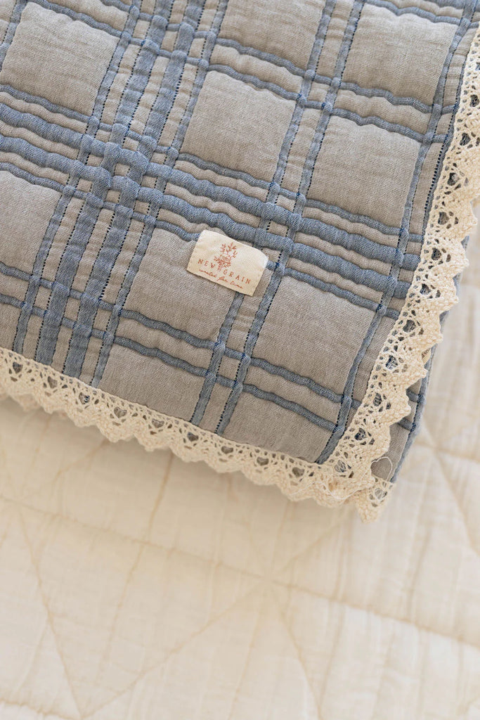 The Organic Cotton Baby Quilt - French Plaid, Bluebelle features a folded light blue and gray checkered pattern with cream lace trim and "New Roman" label, made in Portugal, measuring 30x40 inches, shown atop a cream quilt.