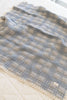 The Organic Cotton Baby Quilt - French Plaid, Bluebelle, featuring a soft blue and gray plaid pattern with lace trim, is displayed on a light-colored textured surface. Made in Portugal.