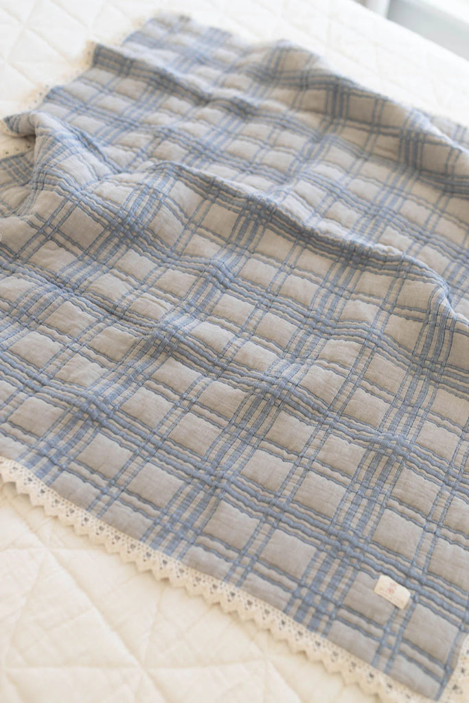 The Organic Cotton Baby Quilt - French Plaid, Bluebelle, featuring a soft blue and gray plaid pattern with lace trim, is displayed on a light-colored textured surface. Made in Portugal.