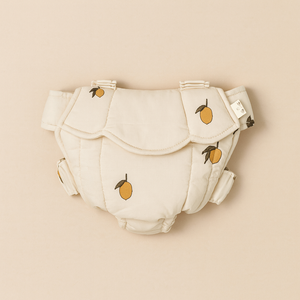 The Konges Sloejd Doll Carrier - Lemon is a soft, cream-colored carrier with embroidered yellow lemons and green leaves. Made from OEKO-TEX certified fabric, it features padded panels and adjustable side straps. Displayed on a beige background.