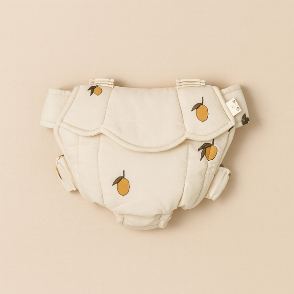 The Konges Sloejd Doll Carrier - Lemon is a soft, cream-colored carrier with embroidered yellow lemons and green leaves. Made from OEKO-TEX certified fabric, it features padded panels and adjustable side straps. Displayed on a beige background.