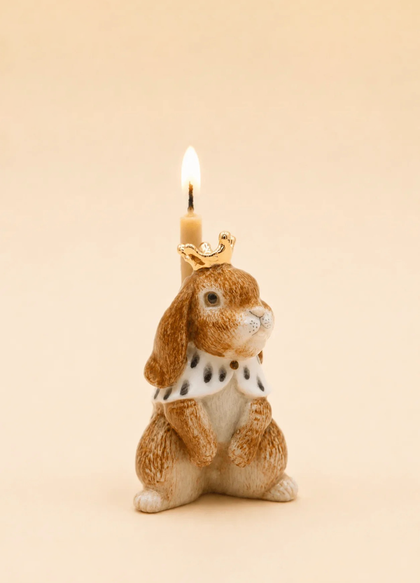 The Highness Hare Cake Topper - Final Sale (ships in one week) features a ceramic bunny with gold accents, a royal cape, and crown, holding a candle—a unique handcrafted centerpiece against a beige background.