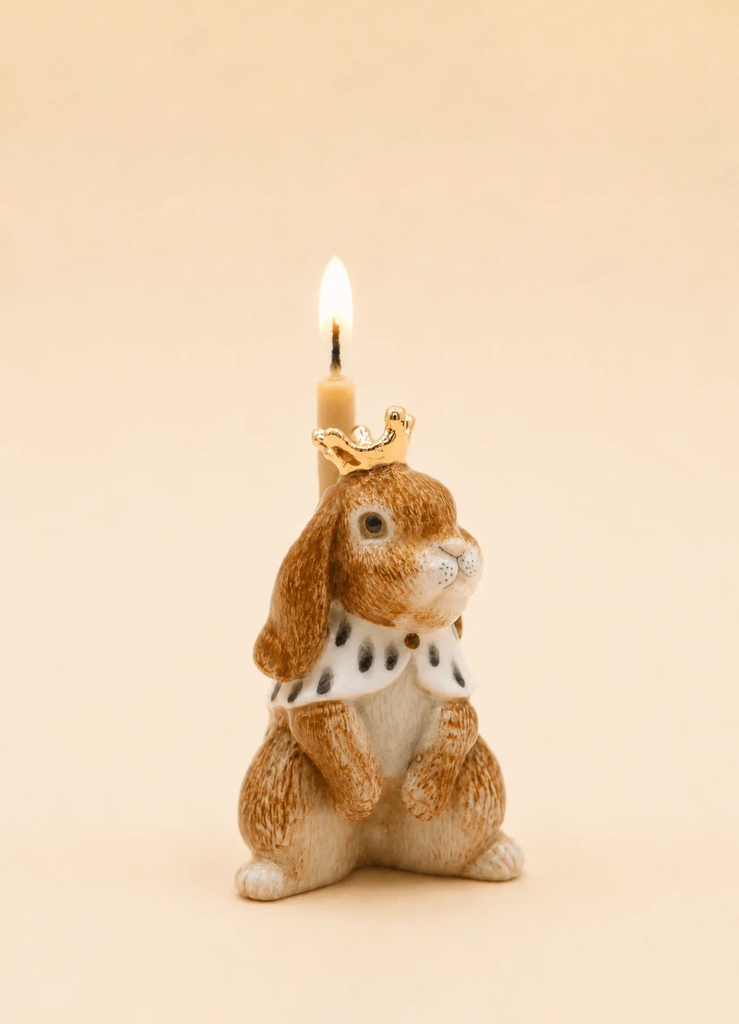 The Highness Hare Cake Topper - Final Sale (ships in one week) features a ceramic bunny with gold accents, a royal cape, and crown, holding a candle—a unique handcrafted centerpiece against a beige background.