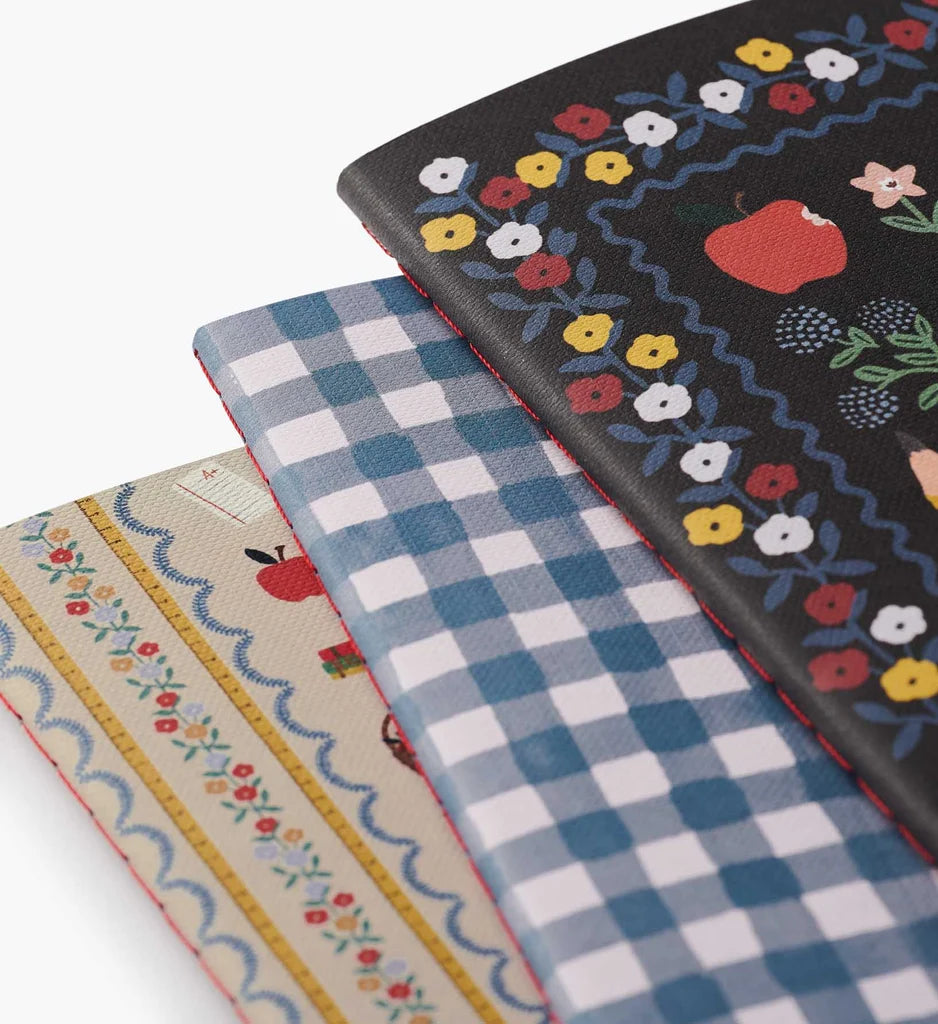A trio from the Assorted Set of 3 Schoolhouse Notebooks is stacked with partial overlap: the top notebook sports a black cover decorated with florals and apples, the middle displays a blue gingham pattern, and the bottom features cream coloring accented by floral and apple motifs. This set is designed with lay-flat binding for comfortable writing and effortless note-taking.