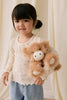 A young girl with dark pigtails, dressed in a cream shirt and light blue jeans, holds Snuggle Bunnies - Hilda The Highland Cow - Caramel plush, her perfect cuddle companion, while standing before light-colored curtains.