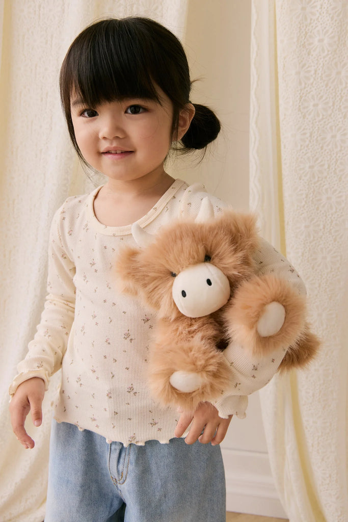 A young girl with dark pigtails, dressed in a cream shirt and light blue jeans, holds Snuggle Bunnies - Hilda The Highland Cow - Caramel plush, her perfect cuddle companion, while standing before light-colored curtains.