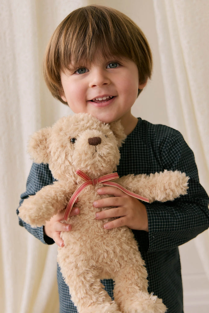 A young child with light brown hair and blue eyes smiles, holding Snuggle Bunnies' Arthur the Bear—Arthur with Red Stripe Bow—in front of a cream-colored curtain.