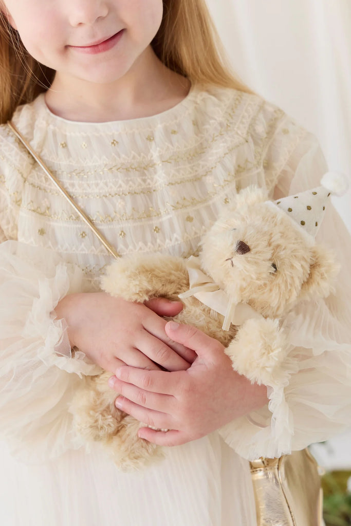 A young girl in a cream-colored dress holds a Snuggle Bunnies - Birthday Arthur - Ivory plush toy, which wears a white party hat with gold polka dots.