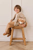 A young child with brown hair, dressed in beige and brown, smiles while hugging Snuggle Bunnies - Hilda The Highland Cow - Caramel plush on a wooden stool, posed in front of a white wall with decorative trim.