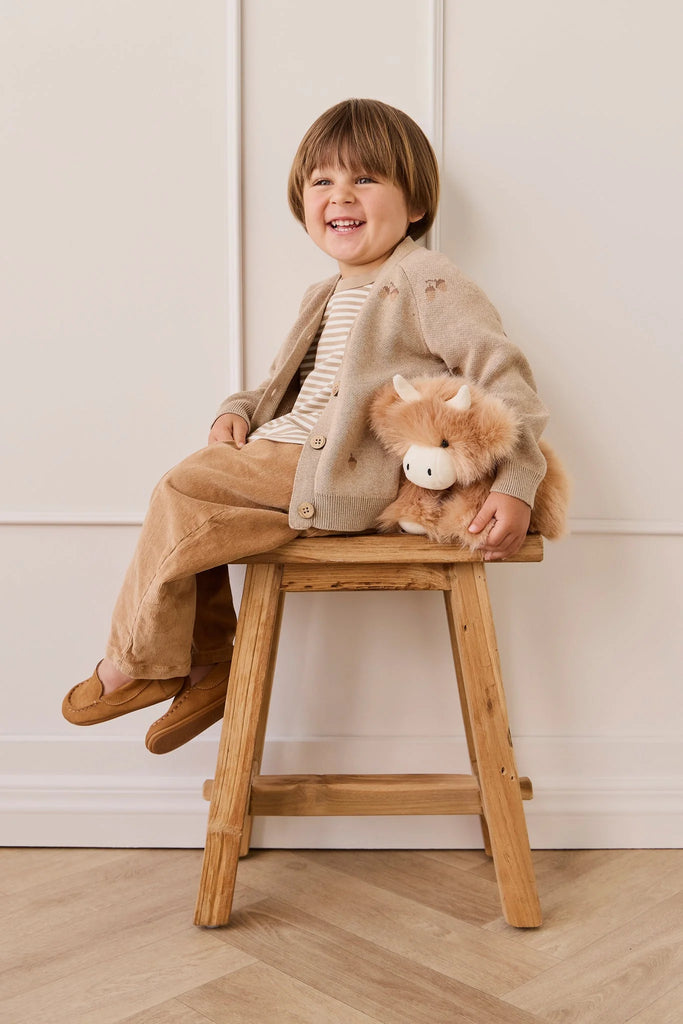 A young child with brown hair, dressed in beige and brown, smiles while hugging Snuggle Bunnies - Hilda The Highland Cow - Caramel plush on a wooden stool, posed in front of a white wall with decorative trim.
