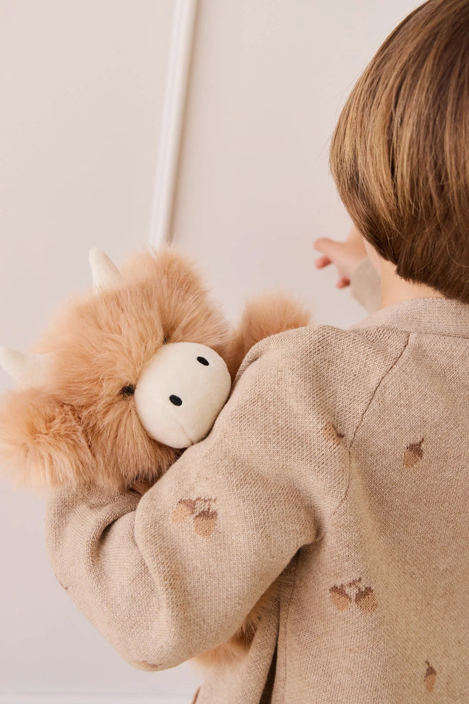 A child with light brown hair, seen from behind, cuddles Snuggle Bunnies - Hilda The Highland Cow - Caramel, a fluffy tan plush with white horns and a cream face in a beige sweater with brown spots—the perfect cuddle companion.