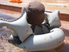 A child wearing a brown sun hat and sitting in an Inflatable Junior Swim Ring - Dinosaurs is enjoying a sunny day in the water. The floatation device, designed for children aged 3-6 years, has two armholes and is decorated with small patterns. The water around the child is clear and shallow.