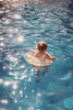A toddler with blond hair is floating on a pool in an Inflatable Junior Swim Ring - Clear Glitter Hearts under sunny skies, with the water sparkling around them.