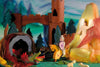 An intricately crafted scene features an Ostheimer Red Knight riding a yellow horse, bravely facing a green dragon emerging from a cave. Behind the knight, an angelic figure stands in front of a glowing yellow and orange backdrop. In the background, wooden castle towers and trees elevate this imaginative play with Ostheimer toys.