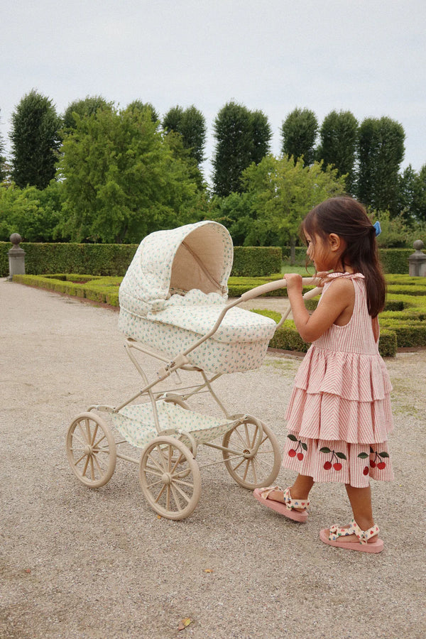 In a picturesque garden lined with manicured hedges and lush trees, a young girl in a pink dress adorned with cherry patterns elegantly pushes the Fleur Coloré doll pram, highlighting its vintage charm crafted from recycled materials along the gravel path.