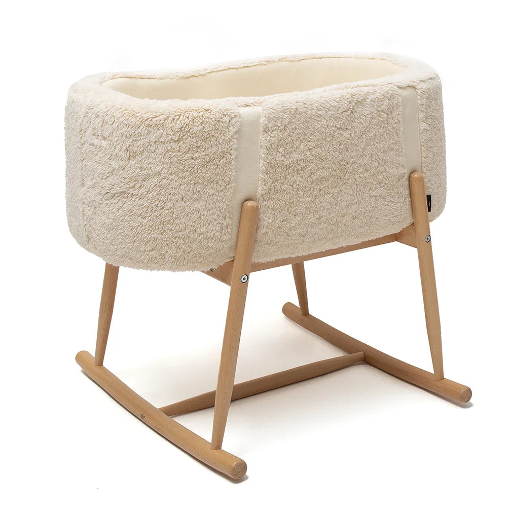 The Charlie Crane KUKO 'Fur' Moses Basket features soft, cream-colored fluffy sides and light wooden rocking legs, creating a modern and elegant look, presented here against a white background.