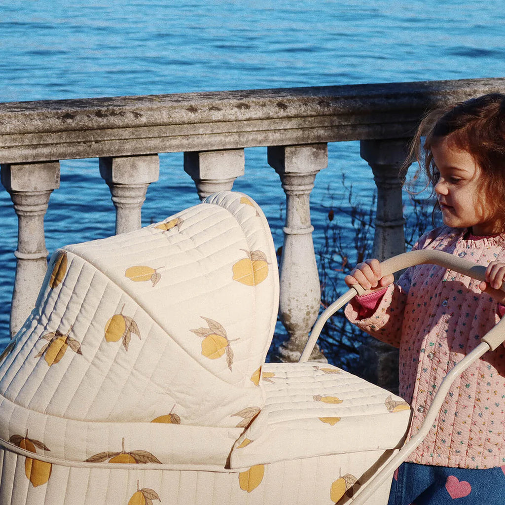 A child in a pink sweater joyfully pushes the Doll Pram - Grand Lemon, featuring a vintage-style cream color embellished with a charming lemon pattern. The scenic lakeside backdrop, framed by a stone rail and blue waters, enhances the allure of this delightful moment.