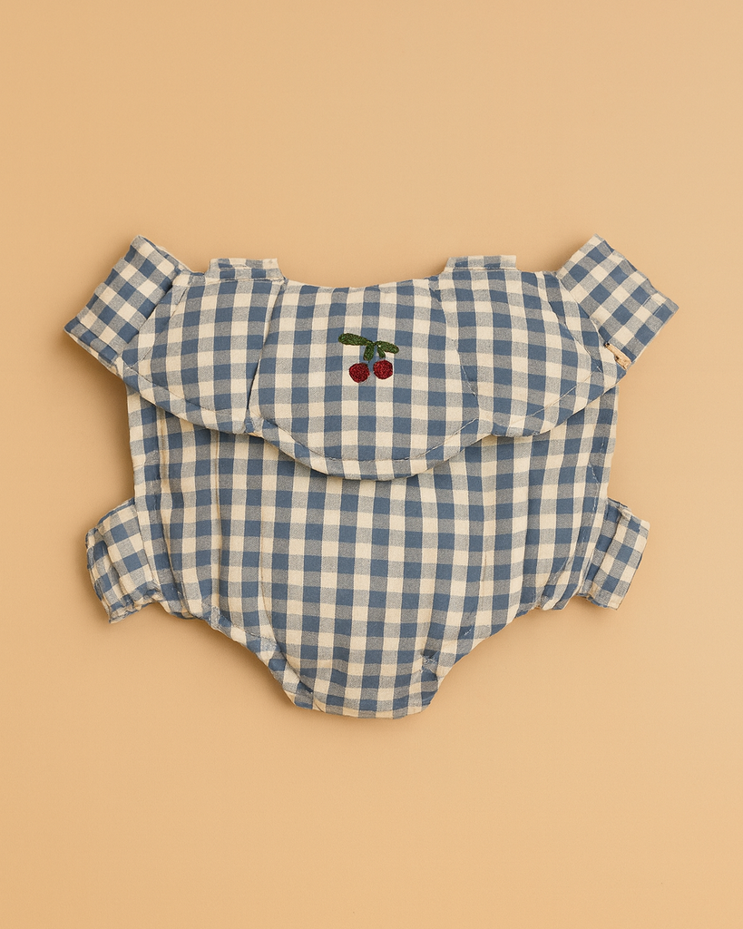 The Konges Sloejd Doll Carrier in Blue Checkers, shown beside a blue and white gingham baby romper with ruffled sleeves and cherry embroidery, makes a charming cotton accessory for your child's doll.