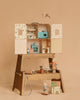 The Margot Step Stool displays a wooden dollhouse with pastel rooms. Mini animal dolls—mice and a bunny—plus miniature furniture, a book, and accessories are set around it on a beige background.