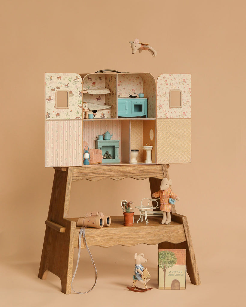 The Margot Step Stool displays a wooden dollhouse with pastel rooms. Mini animal dolls—mice and a bunny—plus miniature furniture, a book, and accessories are set around it on a beige background.
