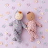 Two Olli Ella Dozy Dinkum - Tootsie Briar baby dolls in floral pajamas lie on a pastel purple surface, surrounded by scattered pastel-colored flower decorations. With their eyes closed and hands joined together, the scene exudes a peaceful and gentle charm.