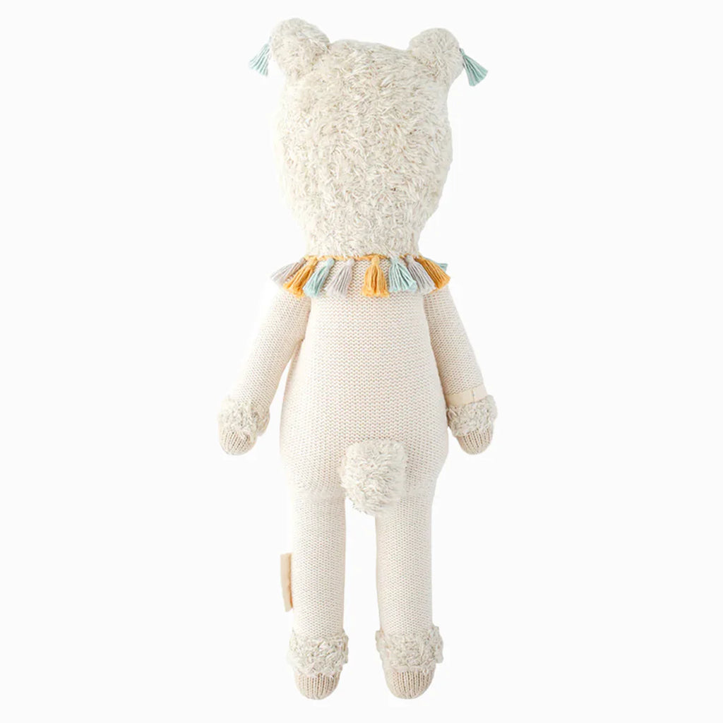 A cream-colored, organic cotton Cuddle + Kind Lucas the Llama plush seen from the back, featuring textured fur on its head, tail, and feet, and wearing a collar with yellow, blue, and white decorative tassels.