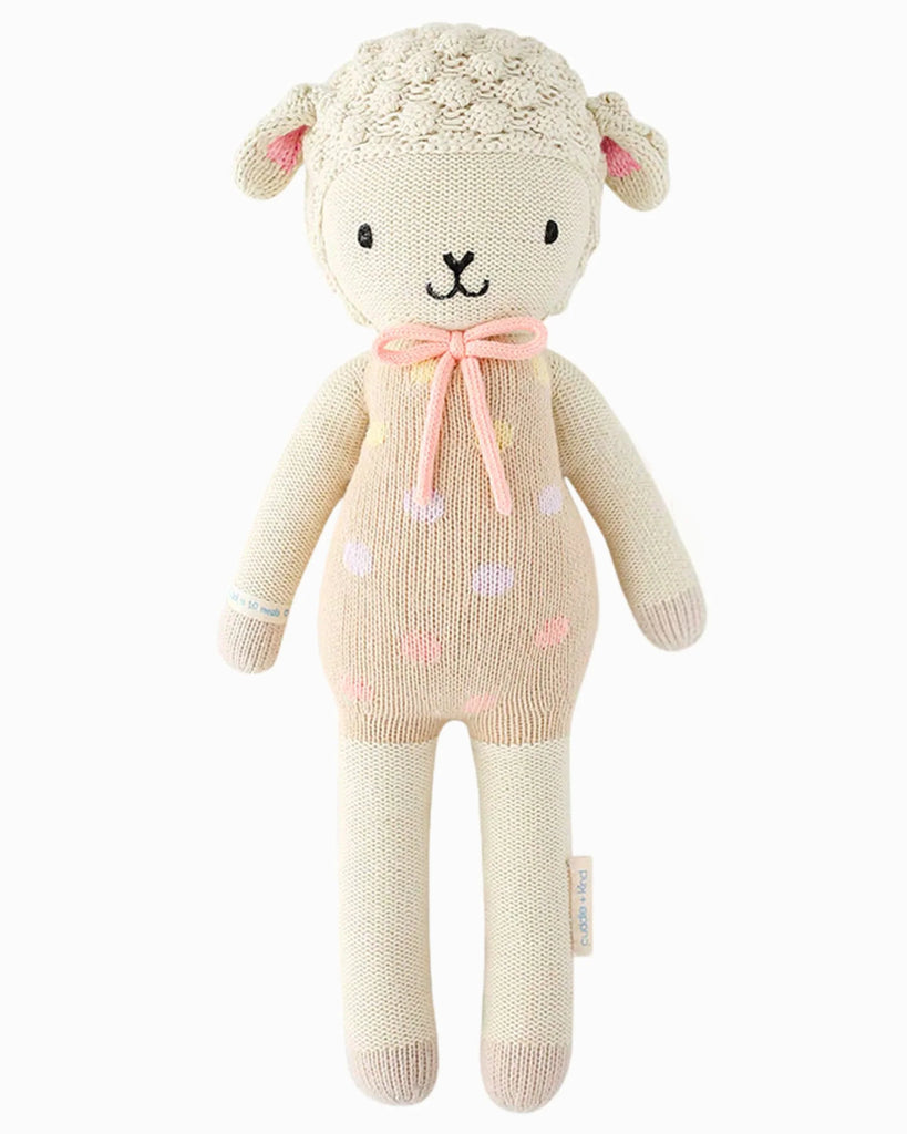 Cuddle + Kind Lucy the Lamb is a hand-knit, hypoallergenic doll with a cream body, beige face, pink cheeks, a pink bow, embroidered details, and pink spots on its sweater.