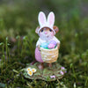 The Wee Forest Folk Tiny Collectible - Hoppy Easter Girl features a hand-sculpted mouse in bunny ears and a pink dress, holding a basket of pastel eggs on a green patch with yellow flowers and moss.