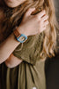 A person with long, wavy hair in an olive green shirt holds their shoulder, wearing a Mini Kyomo Watch - Havana Sky with Japanese movement. The blue and gold watch face is partially visible.