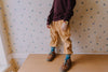 A child stands on a wooden table wearing a maroon sweater, tan pants, blue-green socks, brown shoes, and the Mini Kyomo Watch - Cherry Bomb. Floral wallpaper is in the background; the image is cropped at the shoulders.