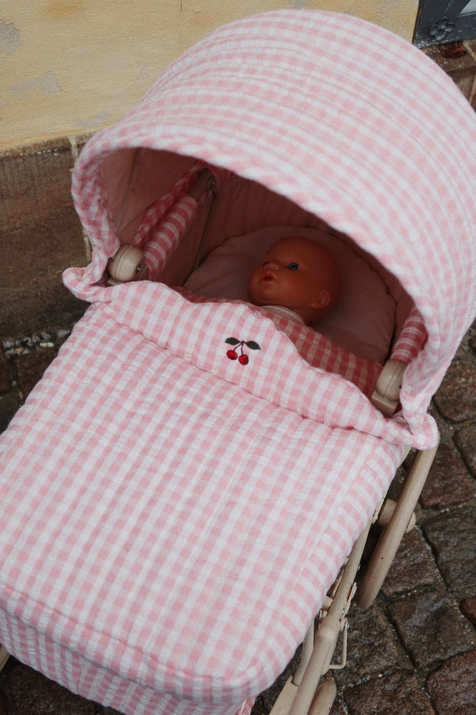 A Konges Sloejd Doll Pram in Powder Pink Checkers cradles a doll, resting on its padded mattress adorned with an embroidered cherry design. The pram is situated on a stone-paved surface adjacent to a plastered wall.